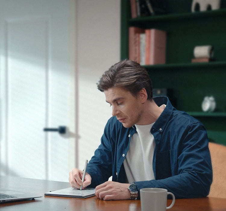 Man in a blue jacket writing in a viwoods AiPaper at a desk with a laptop and mug.